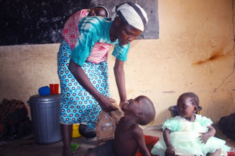 5 year old orphan, Moses Luka, smiles at his aunt as she dresses him in a shirt, as his cousin Ladi looks on. Baby orphan Ibrahim rests on his aunt's back. They arrived in the refugee camp a day ago. January 17, 2015. Photo by Chika Oduah.